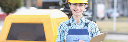 Woman builder in protective helmet holding clipboard with documents in hands on background of...