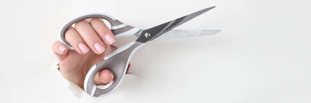 Female hand holds metal scissors in hole made of white paperの写真素材