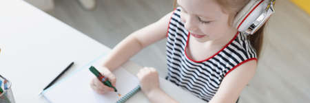 Little girl in headphones sitting in front of laptop and writing in notebook