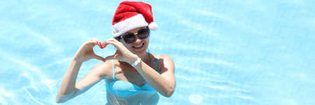 Smiling woman in sunglasses and santa claus hat shows heart gesture while standing in poolの写真素材
