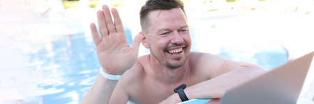 Portrait of emotionally smiling man waving his hand to laptop webcam while in poolの写真素材