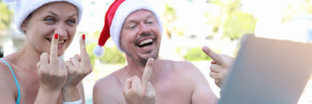 Joyful man and woman in red santa claus hats show gesture of fact in laptop monitor being at resort in poolの写真素材