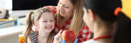 Doctor offering little girl bottle of medicine and red apple in clinicの写真素材