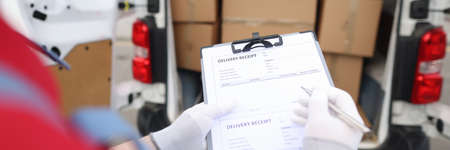 Courier holding in his hands clipboard with delivery receipt and pen near car with cardboard boxes...
