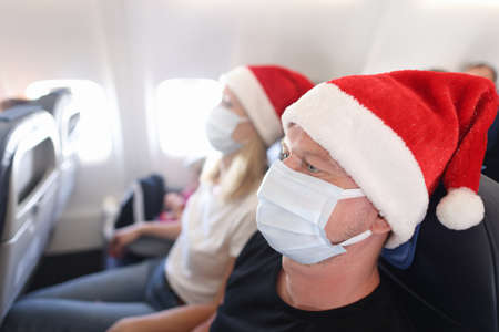 Man and woman wearing red santa claus hats and protective medical masks flying in airplane