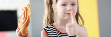 Little girl eating delicious sweet bun and showing thumb up