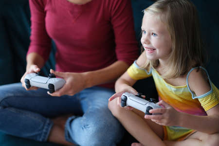 Little girl and mother sitting on couch and holding computer joysticks in their hands