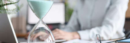 Glass hourglass standing against background of woman working at laptop closeupの写真素材
