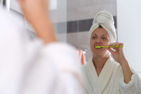 Young woman in bathrobe and with towel on head and brushing teeth in front of mirror in bathroom
