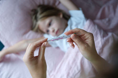 Woman hand holding mercury thermometer with high temperature against background of sick girl closeupの写真素材