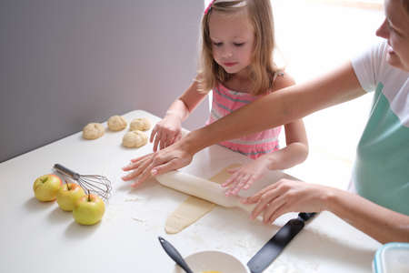 Mom and daughter rolling out flour dough at table in kitchenの写真素材