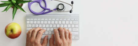 Doctor is typing on keyboard next to stethoscope and appleの写真素材