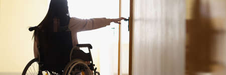 Woman in wheelchair reaches out to open handle of door in dark hallwayの写真素材