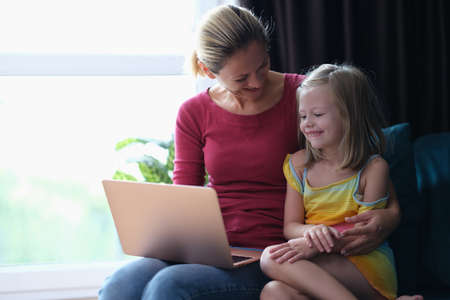 Smiling mom and daughter sitting on couch and looking at laptopの写真素材