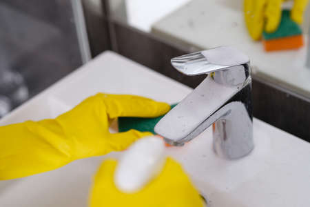 Person in yellow gloves washes sink with washcloth and sprays cleaning foam on faucetの写真素材