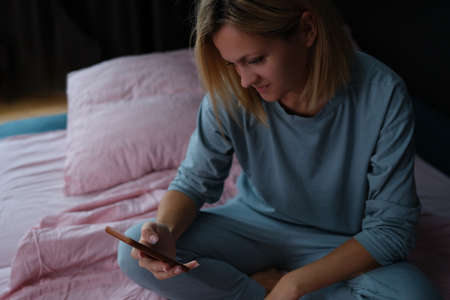 Woman sits on bed and looks into smartphone closeupの写真素材