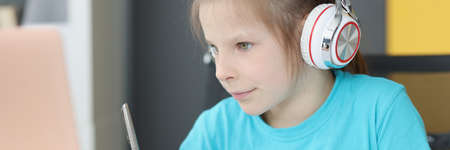 Small preschooler girl in headphones sit at desk study online on laptop