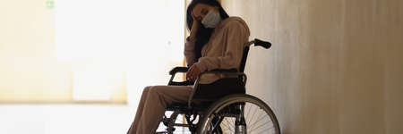 Young disabled woman in protective medical mask sitting in wheelchair in hospital corridorの写真素材
