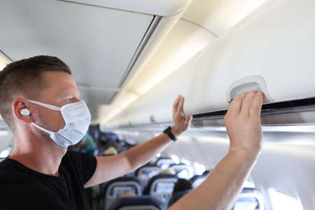 Man wearing protective medical mask opening hand luggage rack on airplaneの写真素材