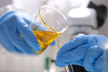 Gloved scientist pours yellow oily liquid from flask into test tube.の写真素材
