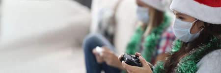Women in protective medical masks and santa claus hats sitting on sofa and holding game console in...