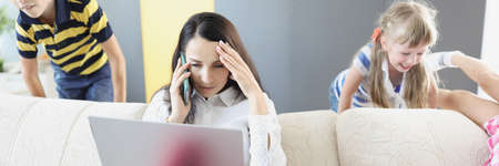 Sad young woman sitting on sofa with laptop and holding her head near children playing at homeの写真素材