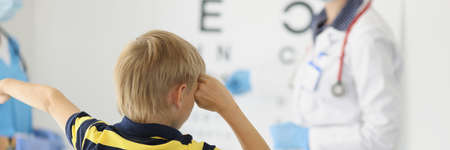 Doctor ophthalmologist n protective mask checking eyesight of little boy at clinic