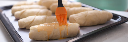 Pastry chef smearing egg yolk on raw bun with silicone brush closeup