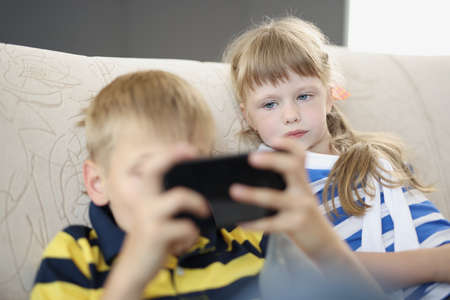 Brother and sister at home on sofa watching on smartphone screenの写真素材