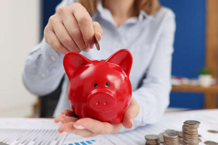 Business woman throwing coins into piggy bank at table with graphs closeupの写真素材