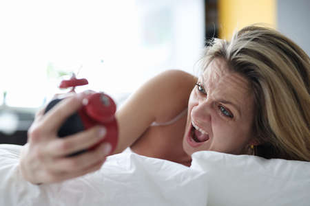 Woman lying in bed with alarm clock in her hands and screaming