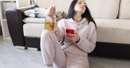 Drunk woman sits on floor with alcohol and writes sms in smartphoneの写真素材