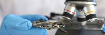 Engineer in glove holds microcircuit against background of microscope