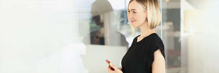 Attractive young blonde businesswoman in stylish black blouse with smartphone in company officeの写真素材