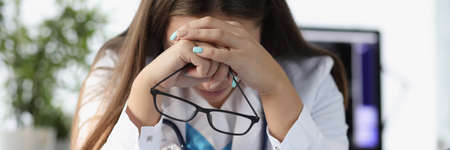 Tired woman doctor sitting at table and holding glasses in clinic
