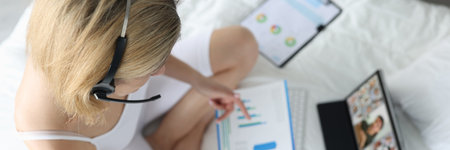 Woman with handsfree sitting on white bed with documents in hands and communicating via video link with colleagues top viewの写真素材