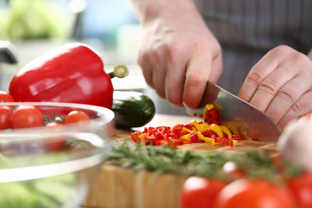 Hands of cook cut yellow and red peppers on cutting board with other vegetables on table in kitchenの写真素材