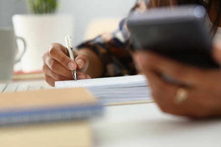 Woman writes in notebook with pen in office and holds smartphoneの写真素材
