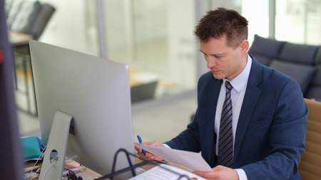 Businessman studying paper document in front of computer in officeの写真素材