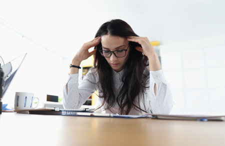 Tired sleepy woman sits at table holds head and rests at the workplace