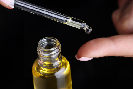 Drop of yellow oil flowing from dropper onto woman finger on black background closeupの写真素材
