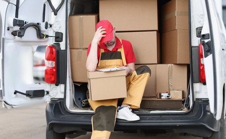 Tired man courier sitting on car with cardboard boxes with documents in his hands