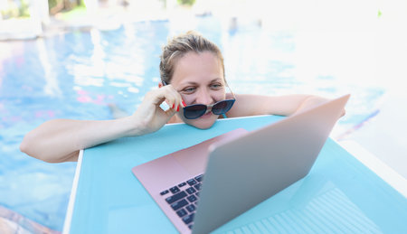 Woman straightening sunglasses and looking into laptop screen in swimming poolの写真素材