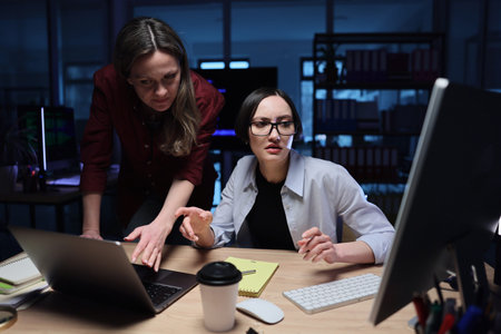 Two women collaborating late at night in a creative workspace filled with technology and...