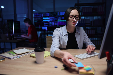 Working late in a modern office, a woman organizes colorful sticky notes while a colleague codes in...