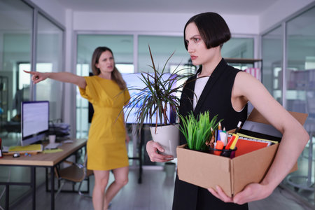 Tension fills the office as a woman leaves with her belongings after a difficult conversation, capturing a moment of uncertainty and changeの写真素材