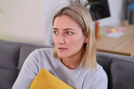 Thoughtful woman gazes out of window while holding a bright yellow pillow in a cozy living room...