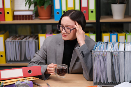 Feeling overwhelmed at work while sipping tea and surrounded by colorful file folders in a bustling...
