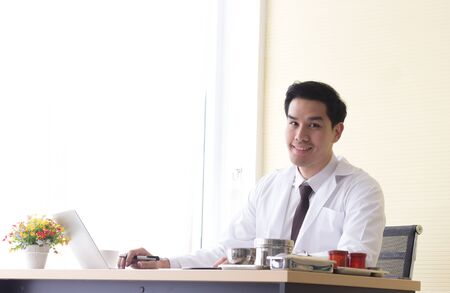 Asian young doctor is smiling on a desk with a notebook and medical equipment in the officeの写真素材