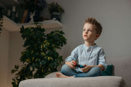 Happy boy playing video games holding game controller sitting on the coach in living room
